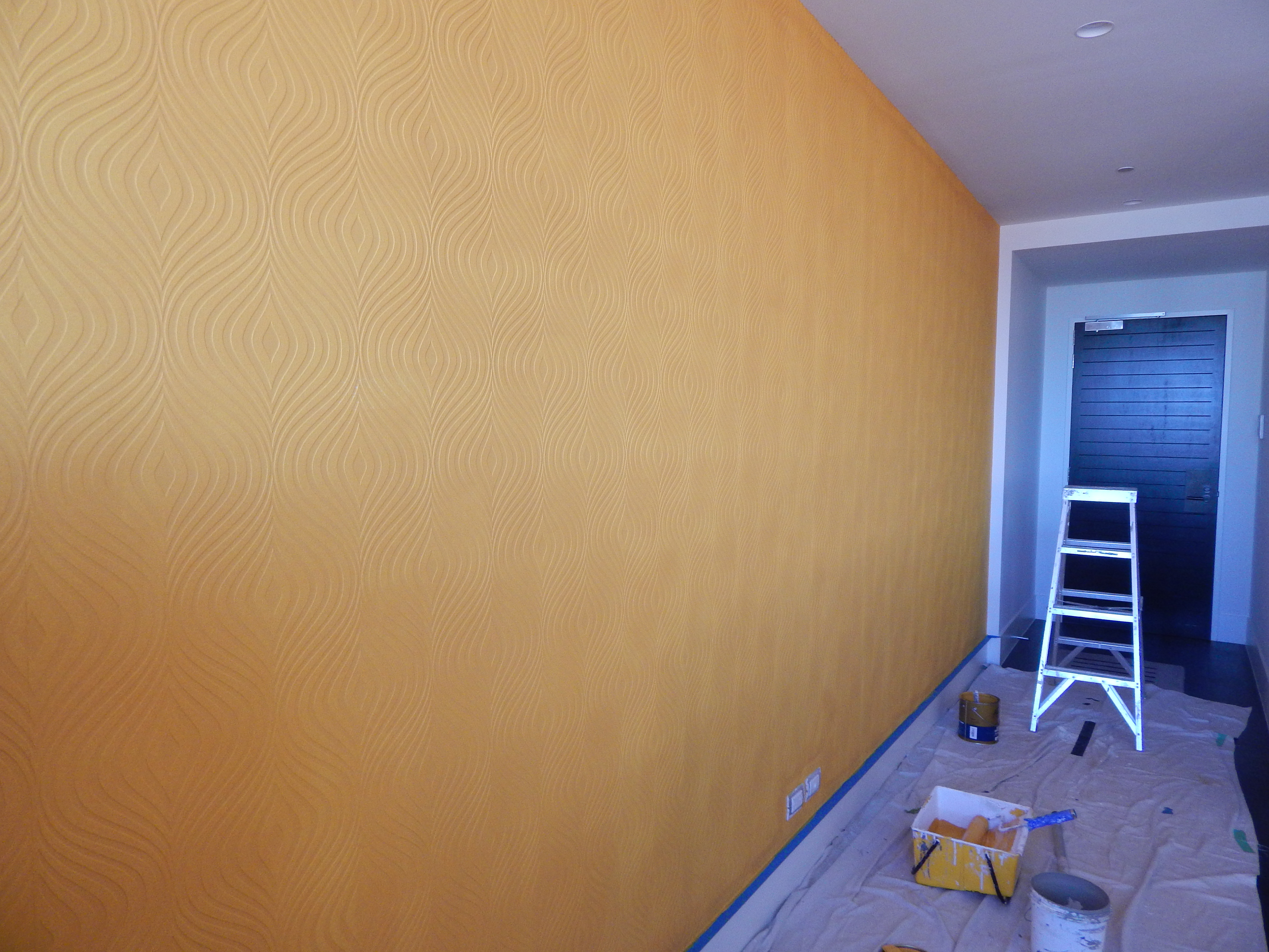 An image of a hallway undergoing home improvement featuring textured orange paint, a white ladder, and paint supplies on a...